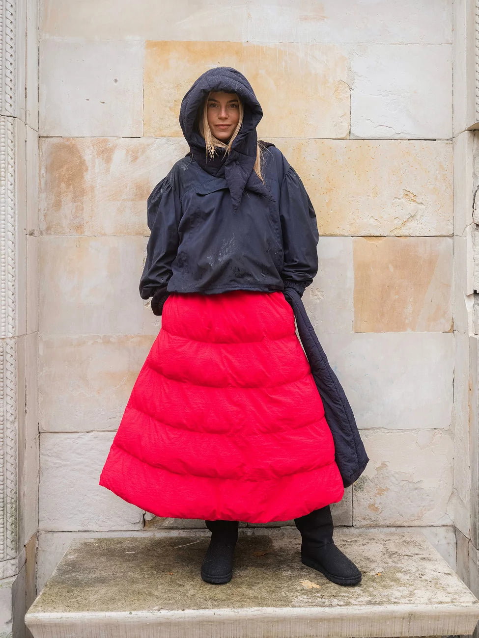 Winter Vegan Skirt Red (limited edition) 1 Person wearing a black hooded jacket and red puffer skirt against a stone wall.