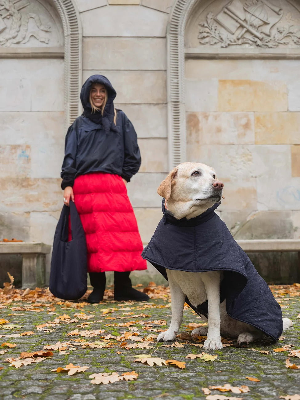 Winter Vegan Skirt Red (limited edition) 5 Woman and dog wearing matching black coats in an urban setting with stone architecture.